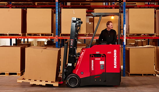 Raymond stand-up counterbalanced forklift transporting pallet in warehouse racking.